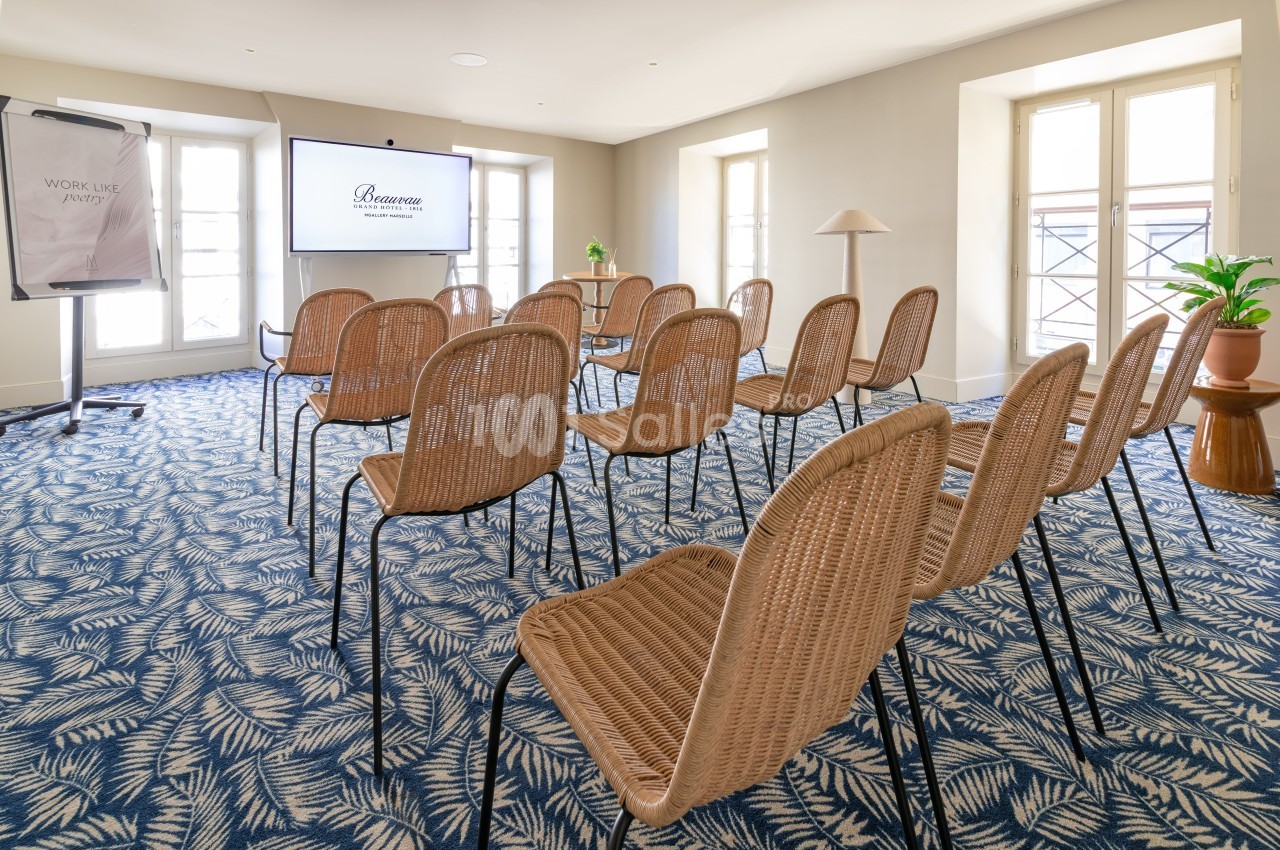 Salle de réunion avec chaises en rotin disposées en U, écran mural, baies vitrées et moquette à motif bleu et beige.