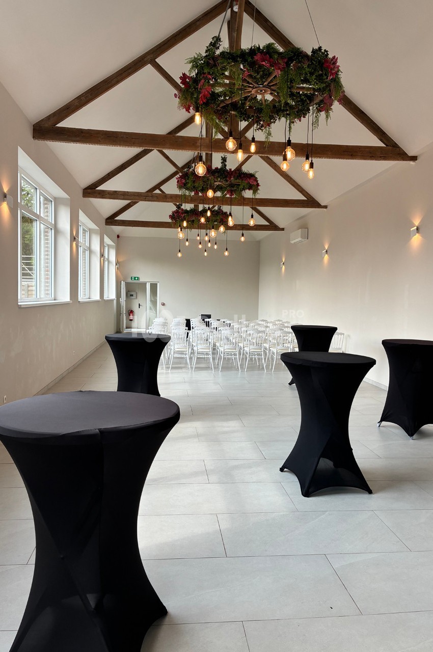 Salle lumineuse avec tables hautes noires, chaises blanches alignées au fond et suspensions décorées de feuillage.