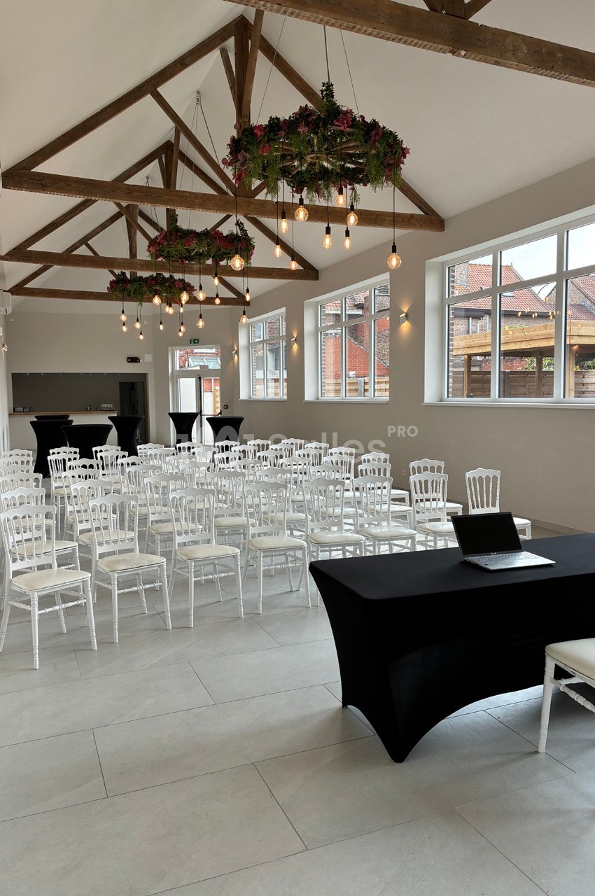 Salle lumineuse avec des chaises blanches alignées, une table noire avec un ordinateur et des décorations suspendues.