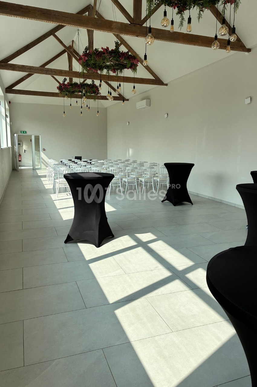 Salle lumineuse avec poutres apparentes, chaises blanches alignées, tables hautes noires et décor floral suspendu.