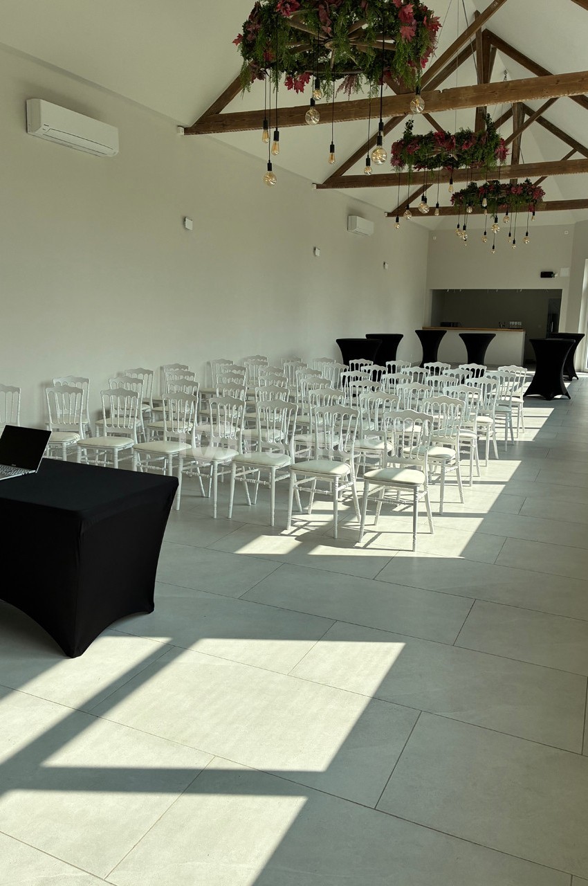 Salle lumineuse aménagée avec des chaises blanches alignées, une table noire et des décorations suspendues.