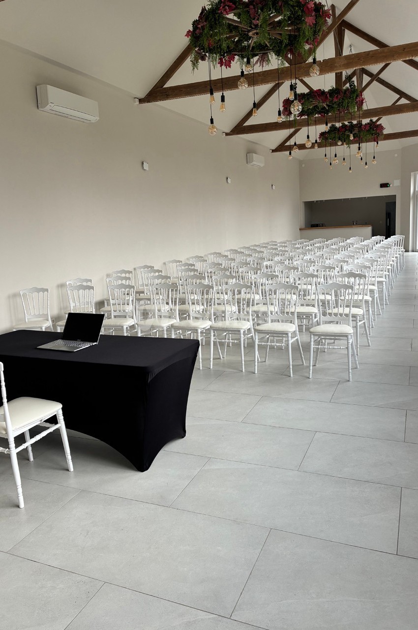 Salle lumineuse aménagée avec des rangées de chaises blanches, une table noire avec un ordinateur portable au premier plan.