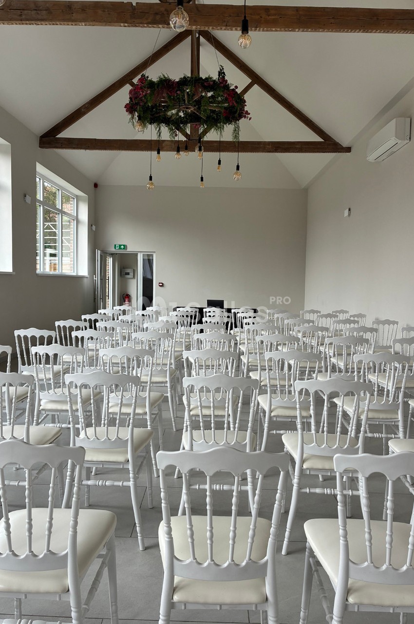 Salle lumineuse avec des chaises blanches alignées, un plafond en poutres apparentes et une décoration florale suspendue.