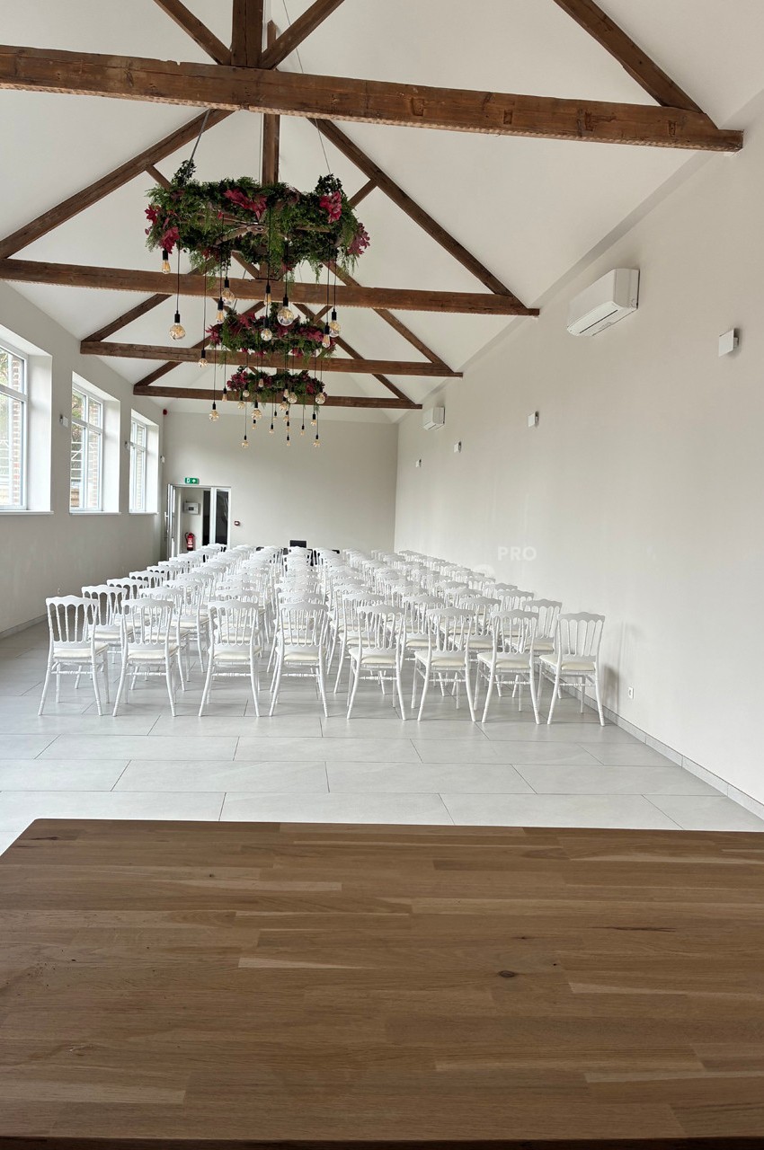 Salle lumineuse avec des chaises blanches alignées, plafond en poutres apparentes et décorations florales suspendues.