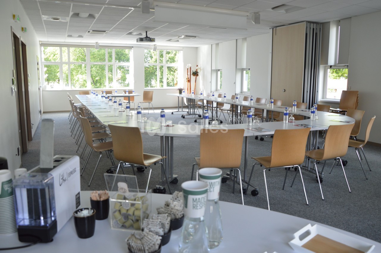 Salle de réunion lumineuse avec tables disposées en U, chaises en bois, et matériel de conférence prêt à l'usage.