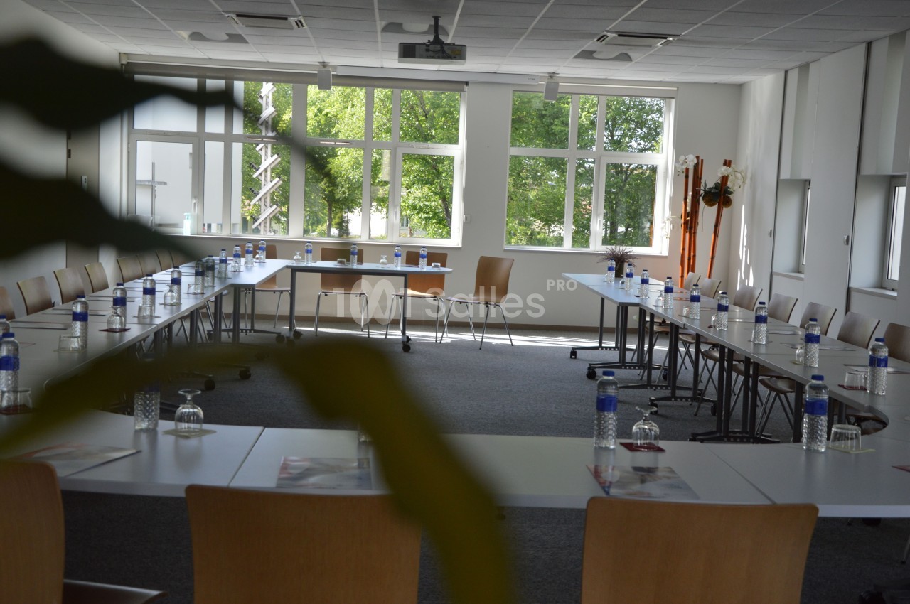 Salle de réunion lumineuse avec des tables disposées en U, des bouteilles d'eau et des verres, vue sur des fenêtres donnant…