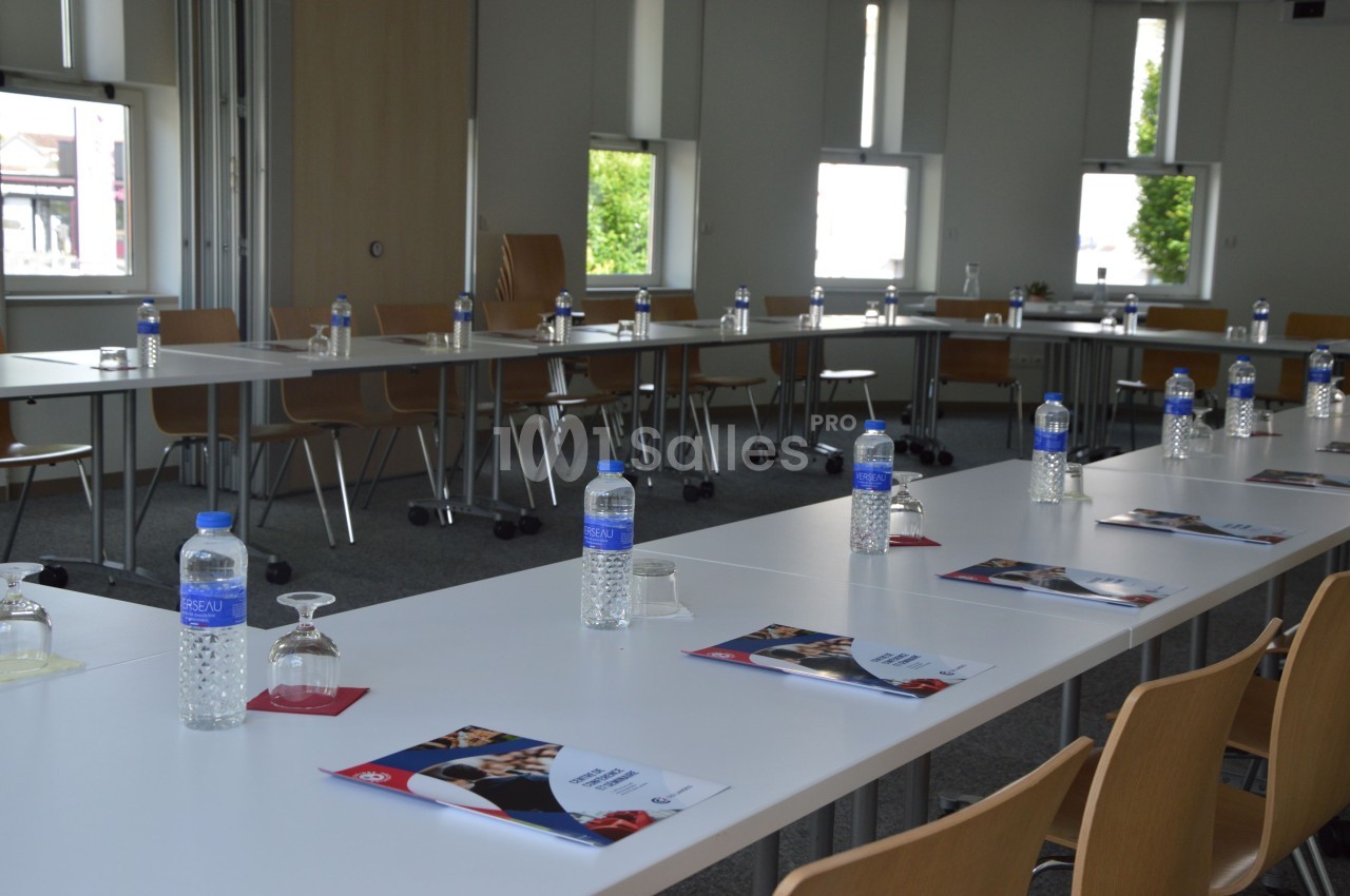 Salle de réunion lumineuse avec des tables en U, des bouteilles d'eau, des verres et des brochures disposés.