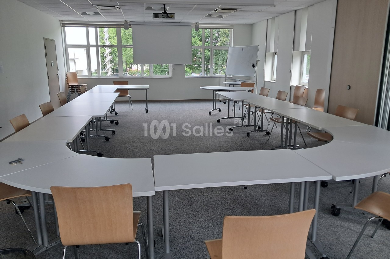 Salle de réunion lumineuse avec des tables disposées en U, des chaises en bois et un tableau blanc au fond.