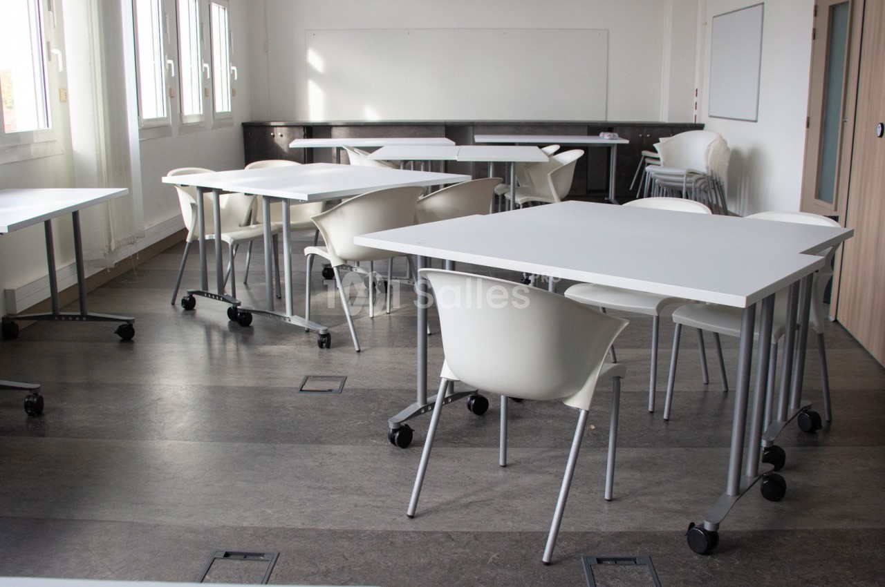 Salle de classe lumineuse avec tables blanches, chaises modernes et tableau blanc au fond.