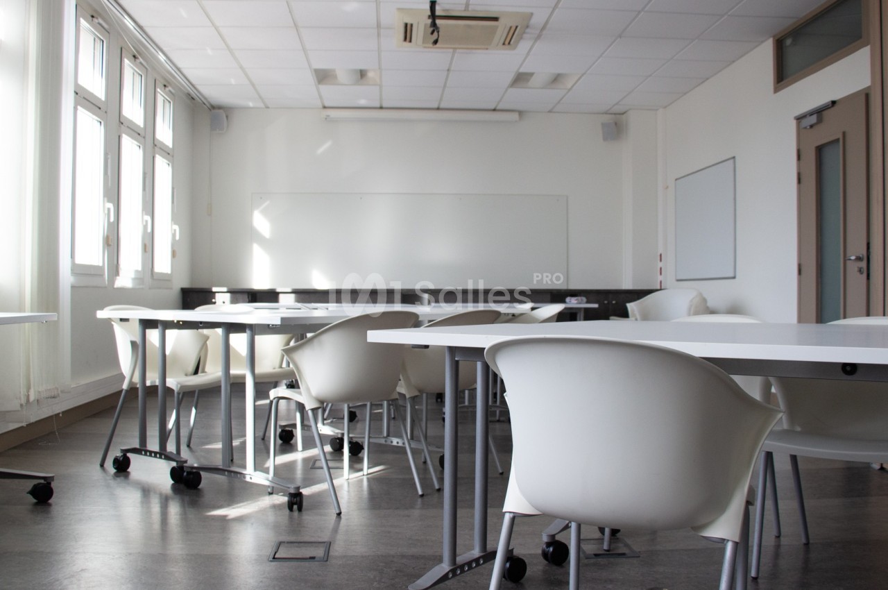 Salle de classe lumineuse avec des tables blanches, des chaises modernes et un tableau blanc au fond.