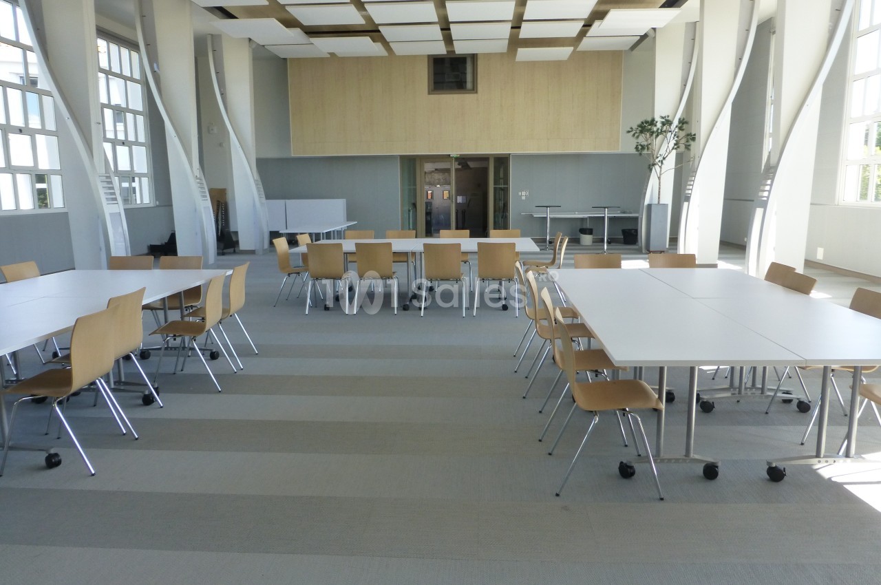 Salle lumineuse avec de grandes fenêtres, tables blanches et chaises en bois disposées pour une réunion ou un travail…