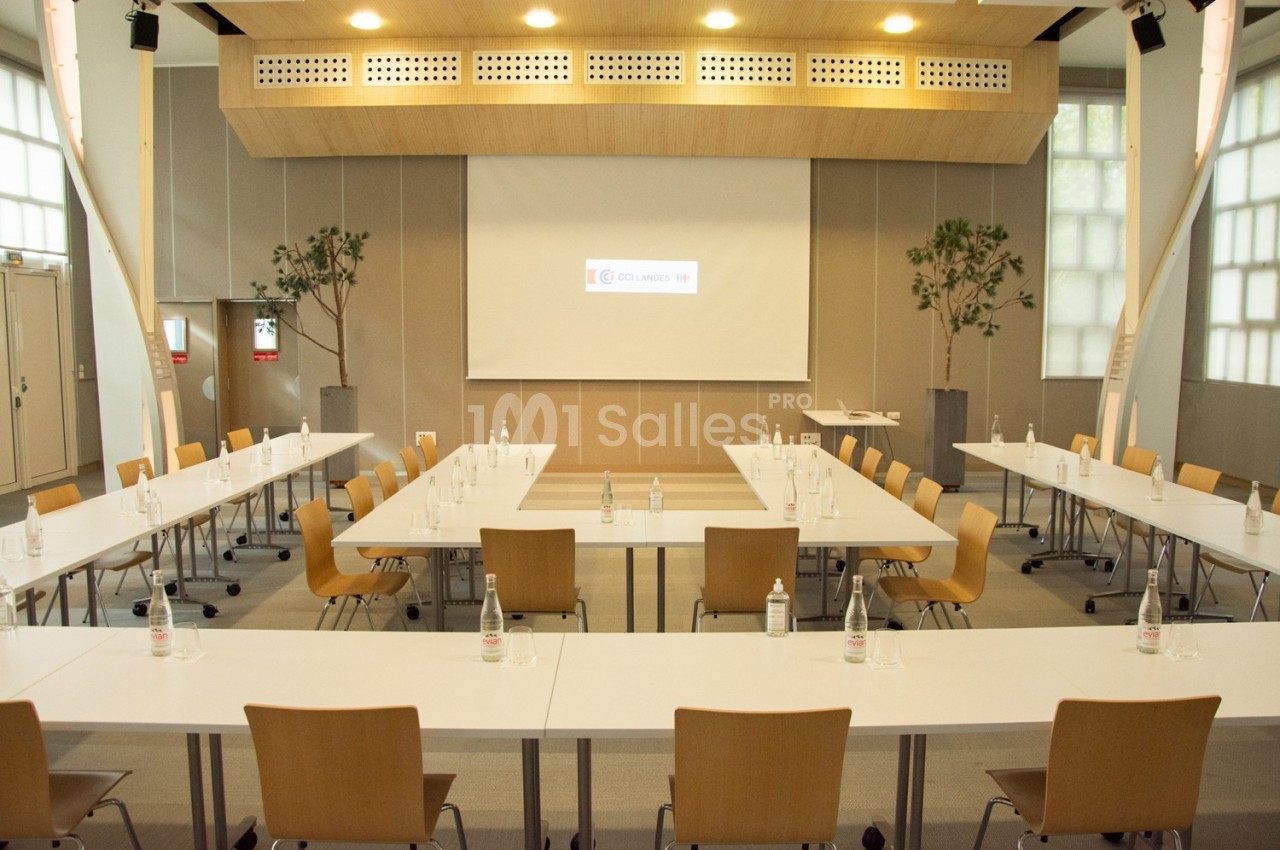 Salle de réunion lumineuse avec tables disposées en U, chaises en bois et écran de projection allumé.