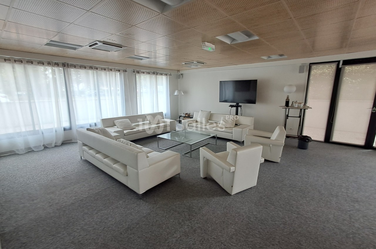 Salle lumineuse avec canapés blancs, table basse en verre, télévision murale et grandes fenêtres avec rideaux.