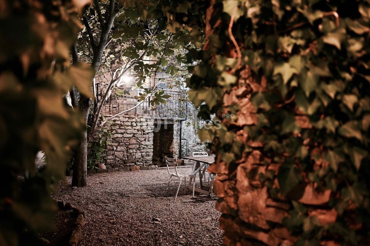 Cour extérieure en soirée, entourée de murs en pierre et de végétation, avec une table et des chaises sous un éclairage doux.