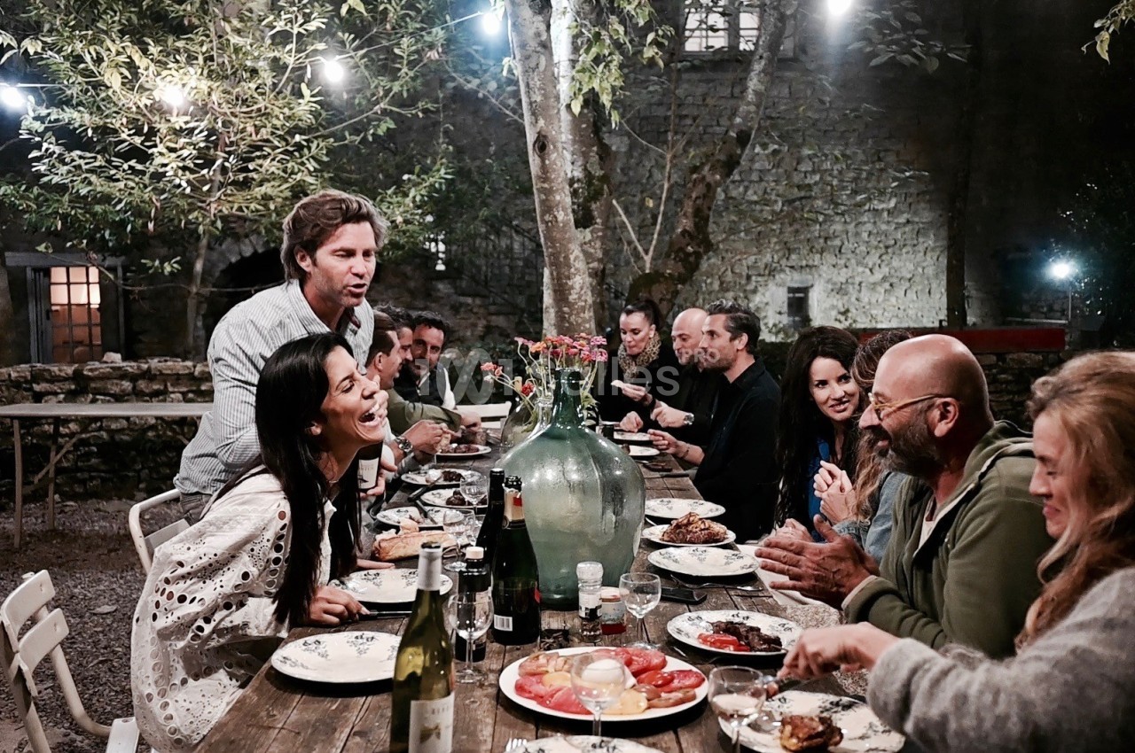 Un groupe de personnes partage un repas convivial en soirée dans un jardin éclairé par des guirlandes lumineuses.