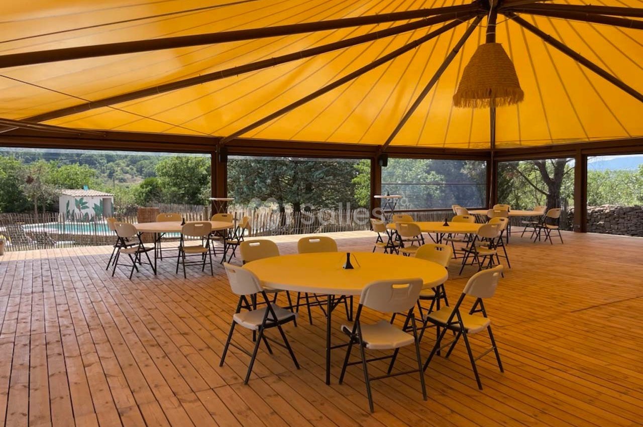 Espace couvert avec tables et chaises sur plancher en bois, entouré de nature et vue sur une piscine.