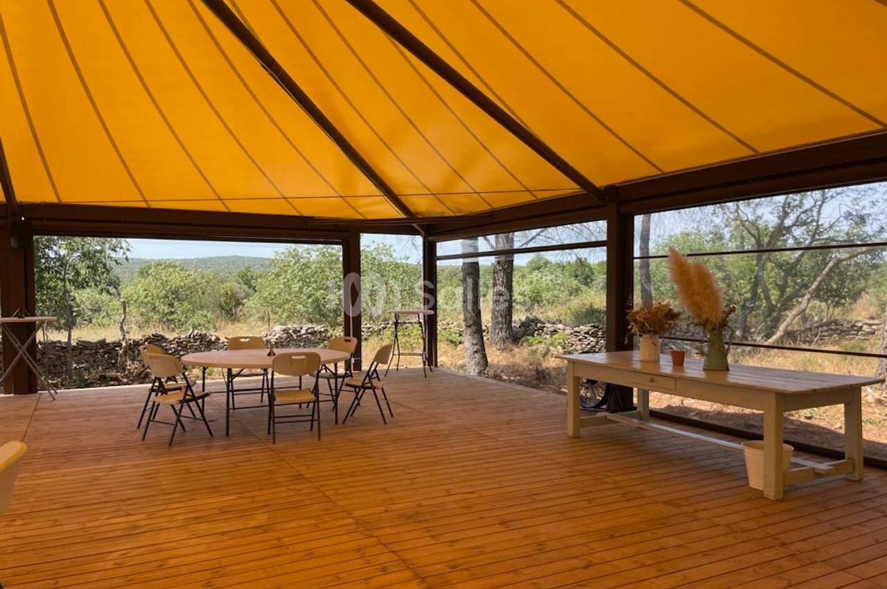 Terrasse en bois couverte d'une toile jaune, avec des tables et chaises, entourée de végétation.
