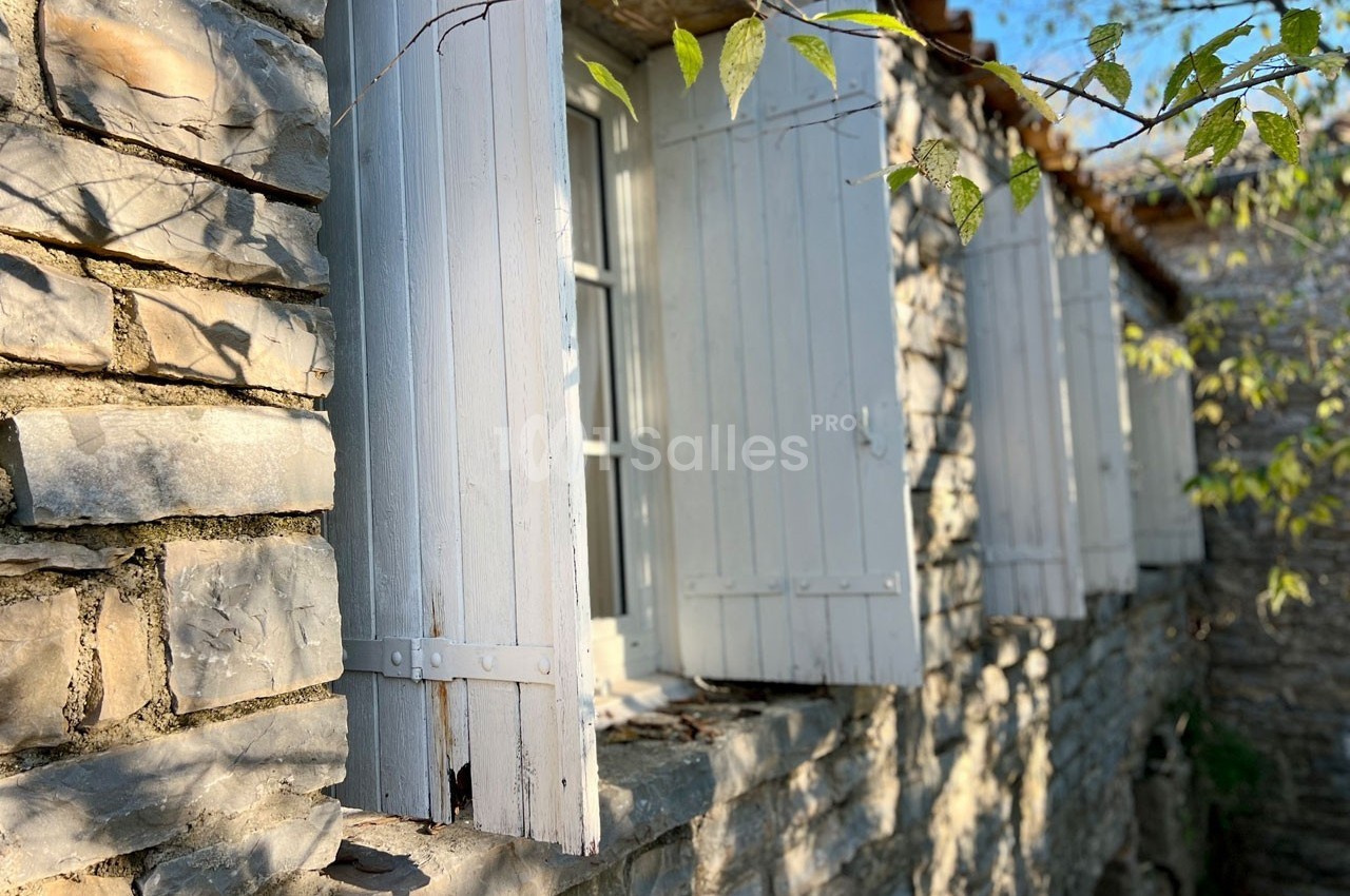 Façade en pierre avec des volets en bois blancs ouverts, éclairée par une lumière naturelle et entourée de végétation.