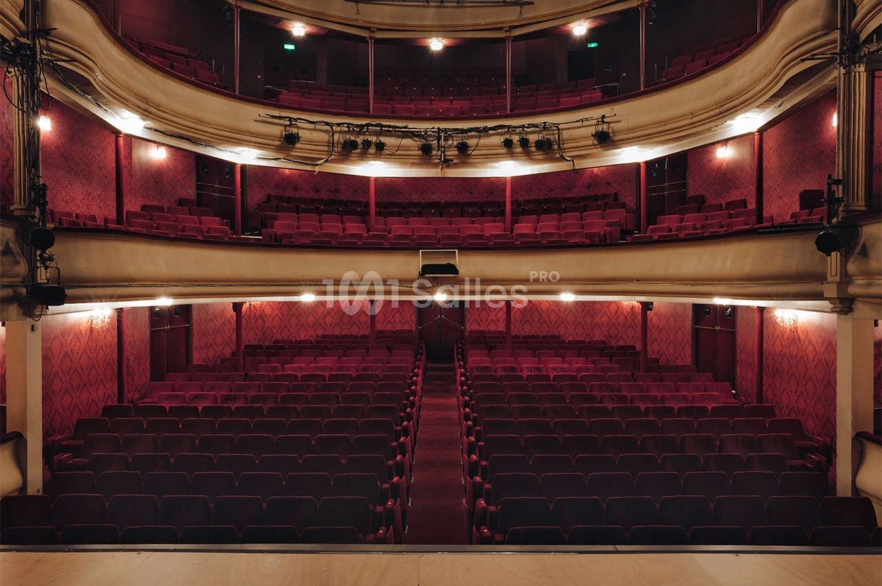 Salle de théâtre vide avec rangées de sièges rouges, balcons ornés et éclairage tamisé.