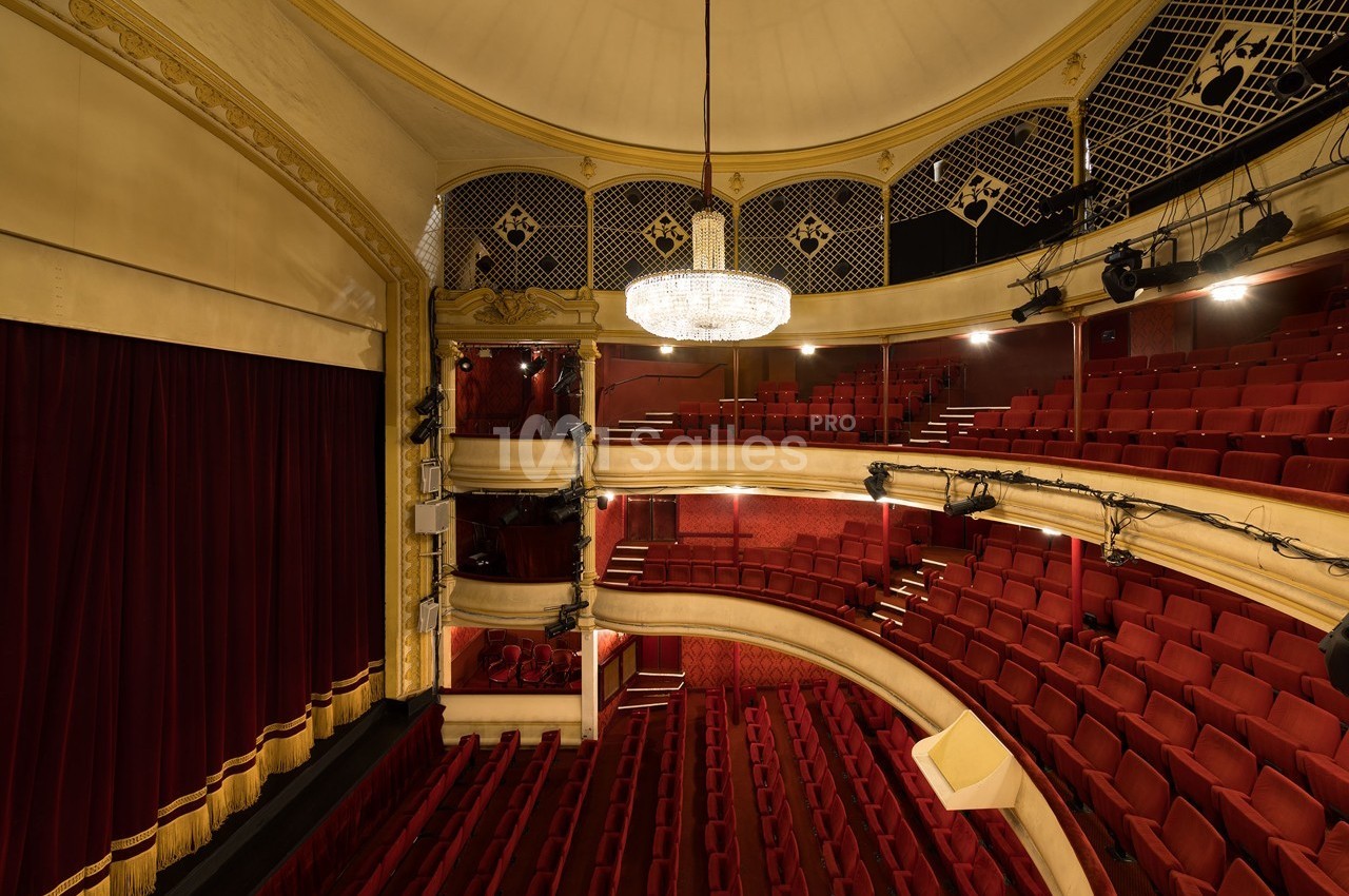 Salle de théâtre avec sièges rouges, balcon, rideaux fermés et grand lustre central sous un plafond voûté.