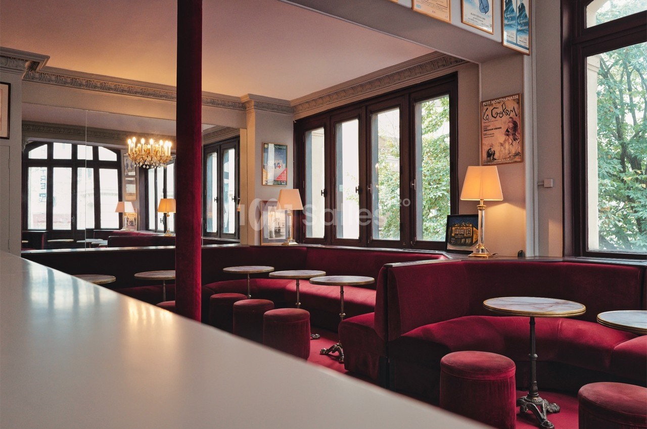 Salle de café élégante avec banquettes rouges, tables rondes en marbre, grandes fenêtres et lustre central.