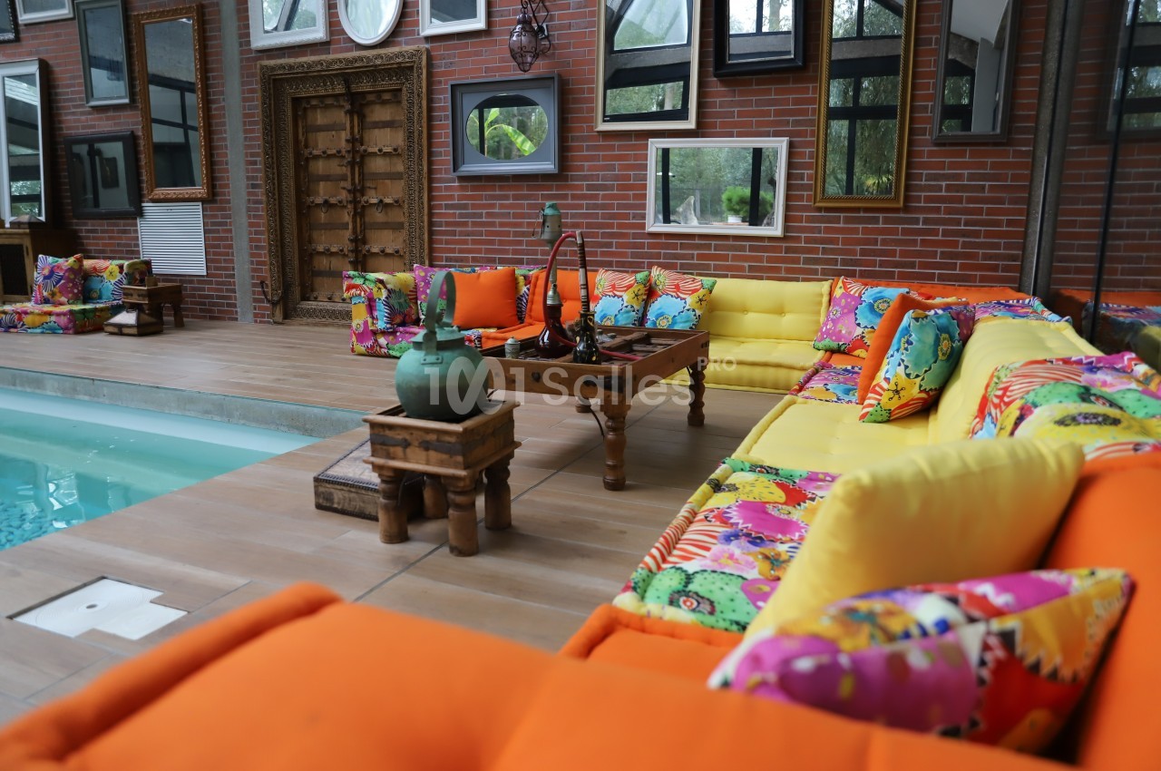Salon extérieur coloré avec canapés orange et jaunes, coussins à motifs vifs, table basse en bois et piscine adjacente.