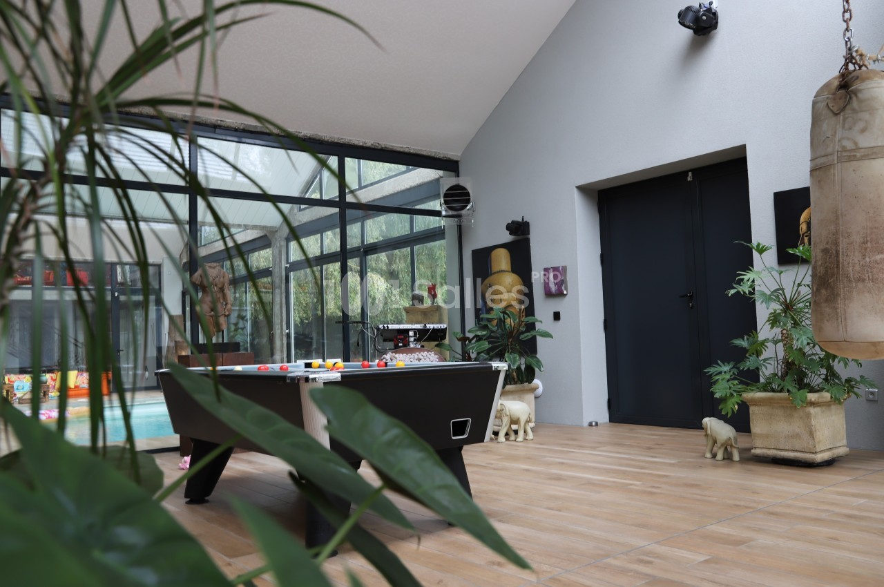 Salle lumineuse avec table de billard, plantes, sac de frappe et vue sur une piscine intérieure à travers des baies vitrées.