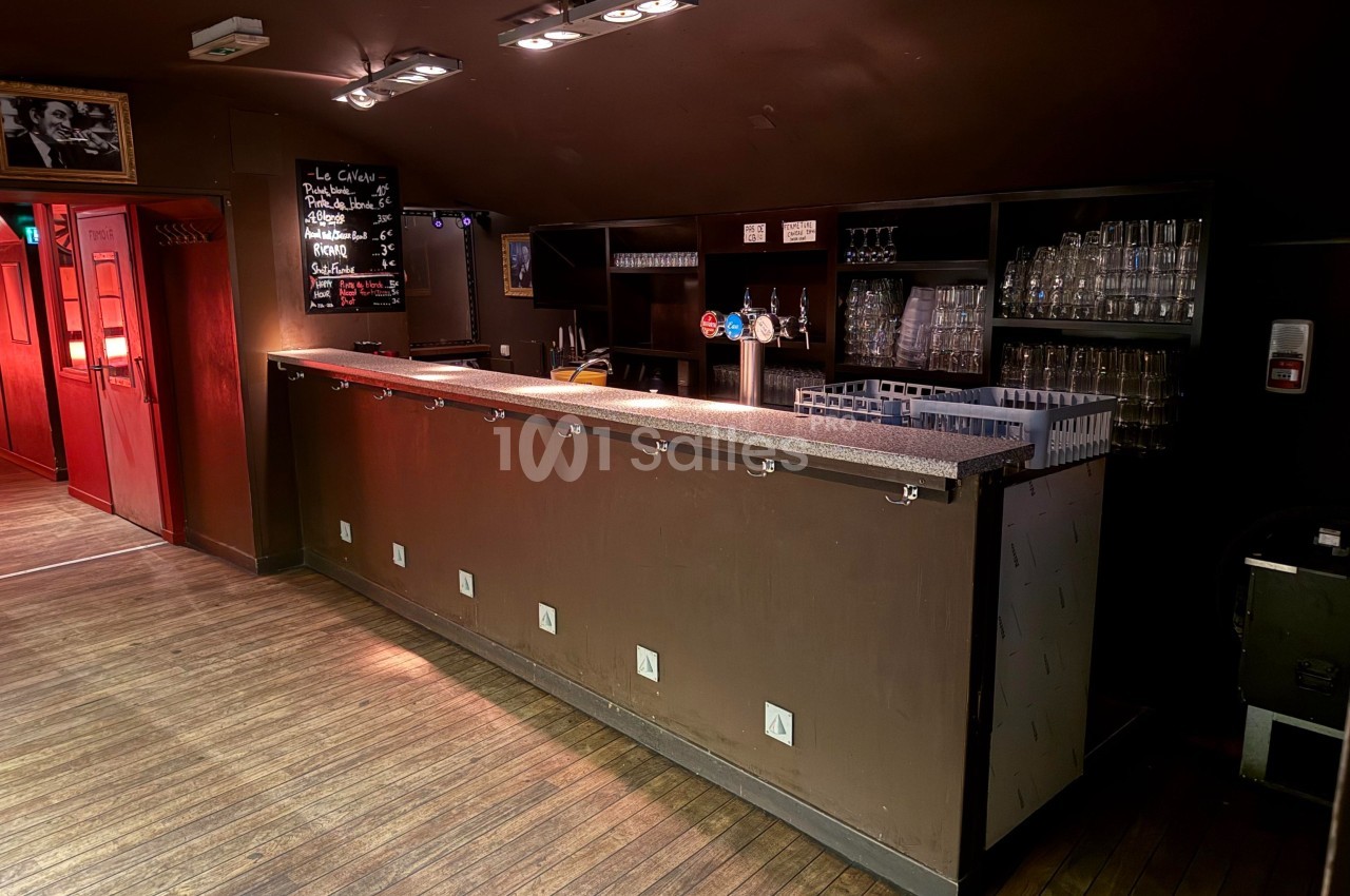 Bar en bois sombre avec comptoir équipé de crochets, étagères à verres et tableau noir affichant le menu.