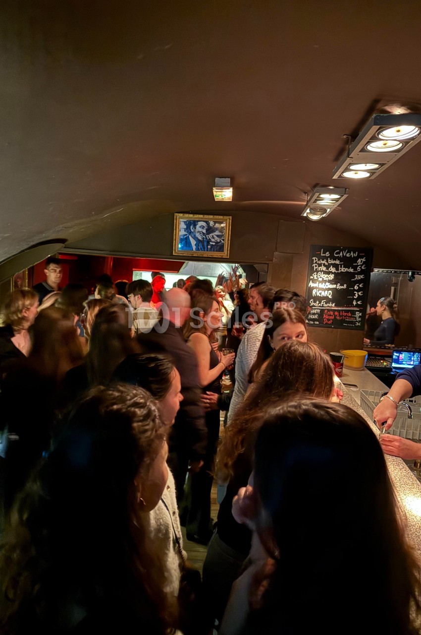 Groupe de personnes discutant et buvant dans un bar animé avec un comptoir éclairé et une ardoise au mur.