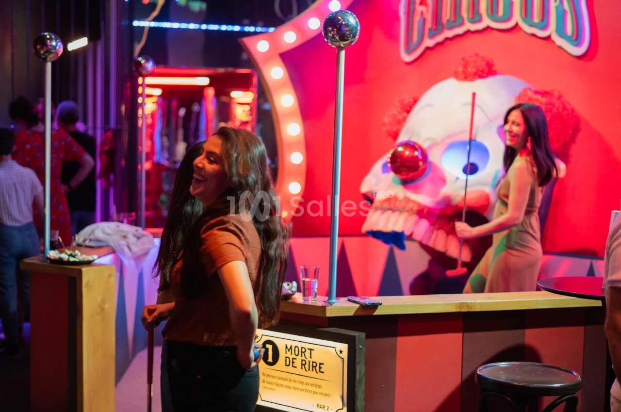 Deux femmes rient près d'un stand de fête foraine avec un décor coloré et une tête de clown en arrière-plan.