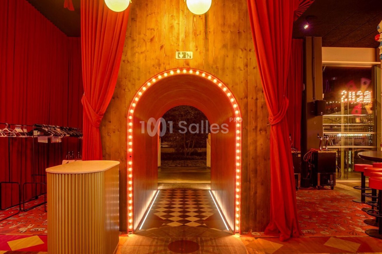 Entrée voûtée éclairée par des lumières, entourée de rideaux rouges, avec un sol à motifs géométriques.