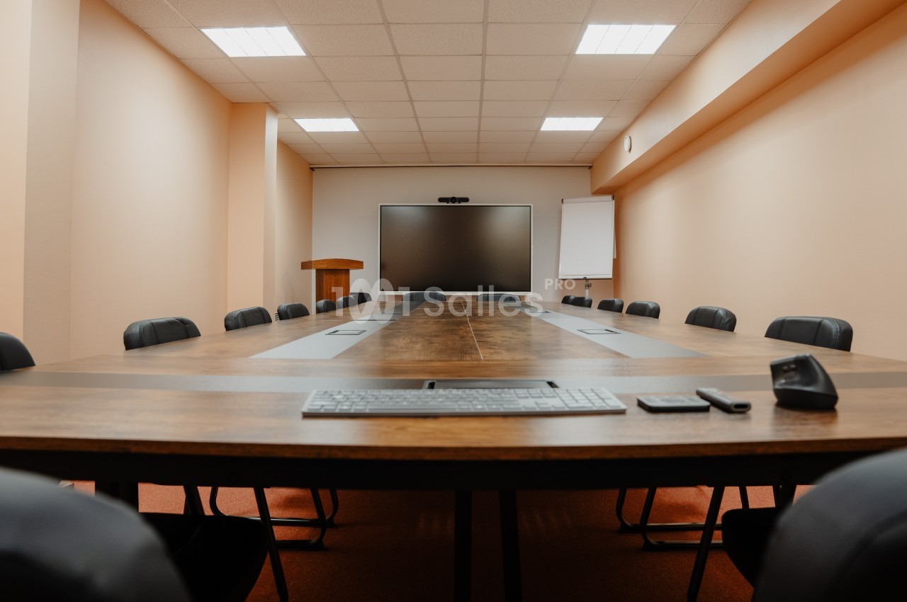 Salle de réunion vide avec une grande table en bois, des chaises noires, un écran mural et un pupitre.