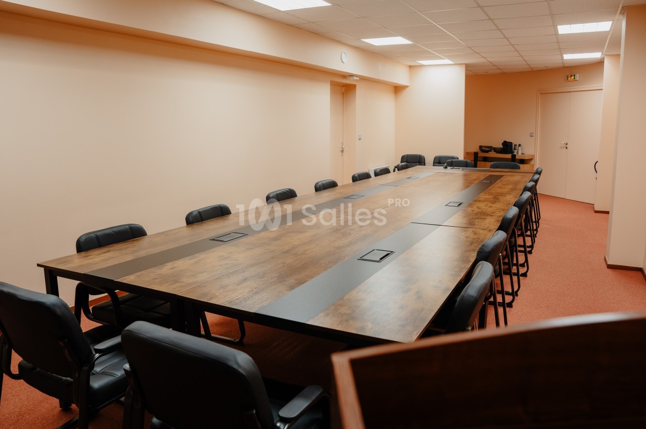 Salle de réunion avec une grande table rectangulaire en bois entourée de chaises noires, éclairée par des lumières au…