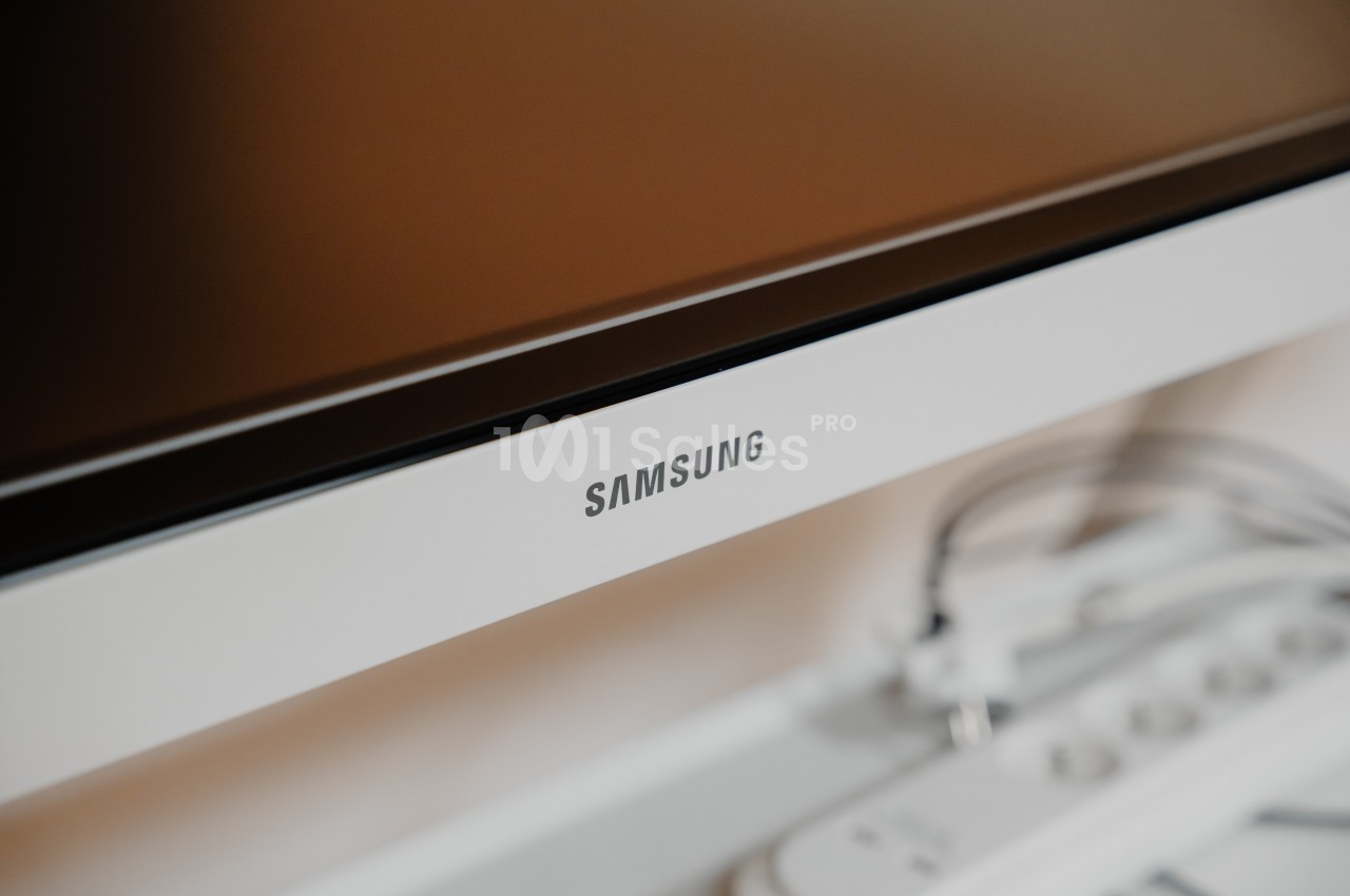 Écran Samsung avec bordure blanche, posé sur un meuble contenant des câbles et des accessoires électroniques.