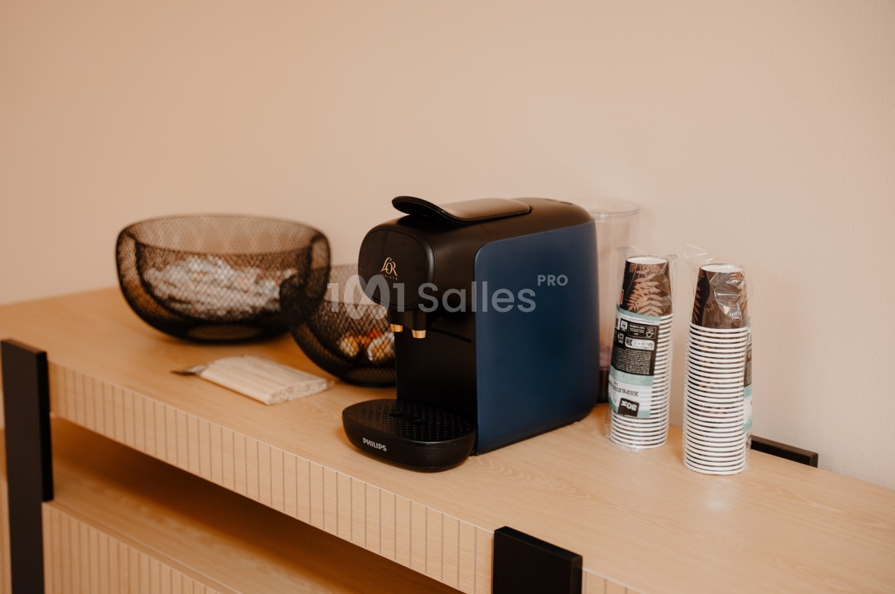 Machine à café noire sur un meuble en bois clair, entourée de gobelets en carton et de paniers en métal.