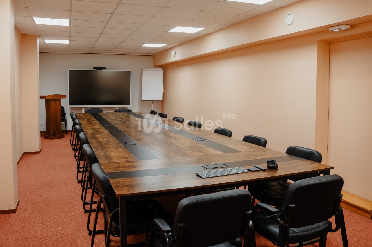 Salle de réunion avec une grande table en bois entourée de chaises noires, écran et tableau blanc en fond.