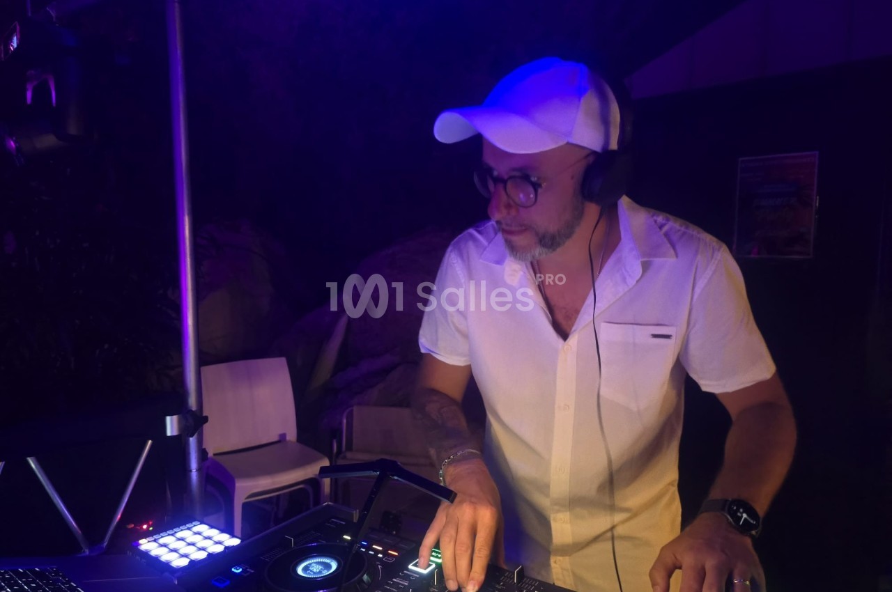 Un DJ en chemise blanche et casquette mixe de la musique sur une table de mixage dans une ambiance nocturne.