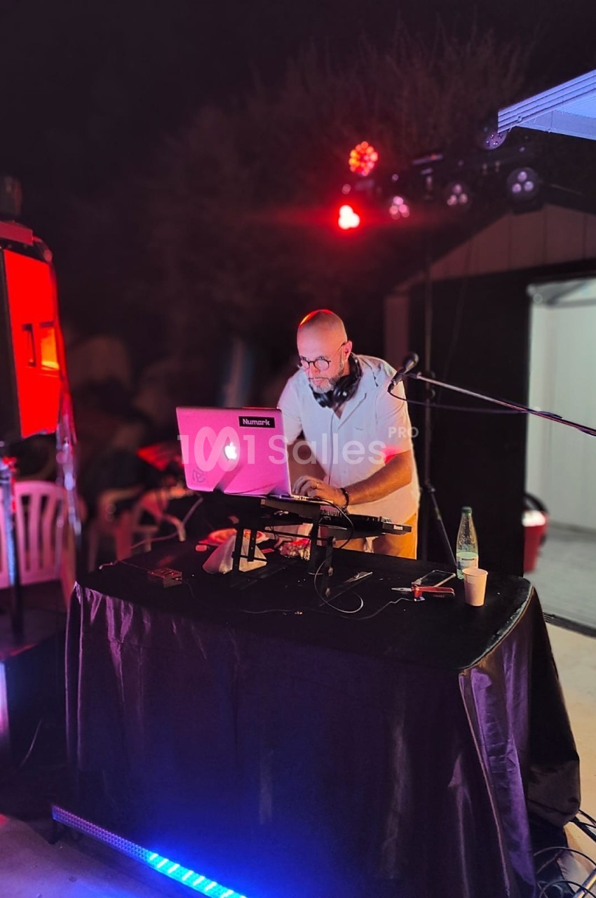 Un DJ mixe de la musique sur une table équipée d'un ordinateur portable et de matériel audio lors d'un événement en soirée.