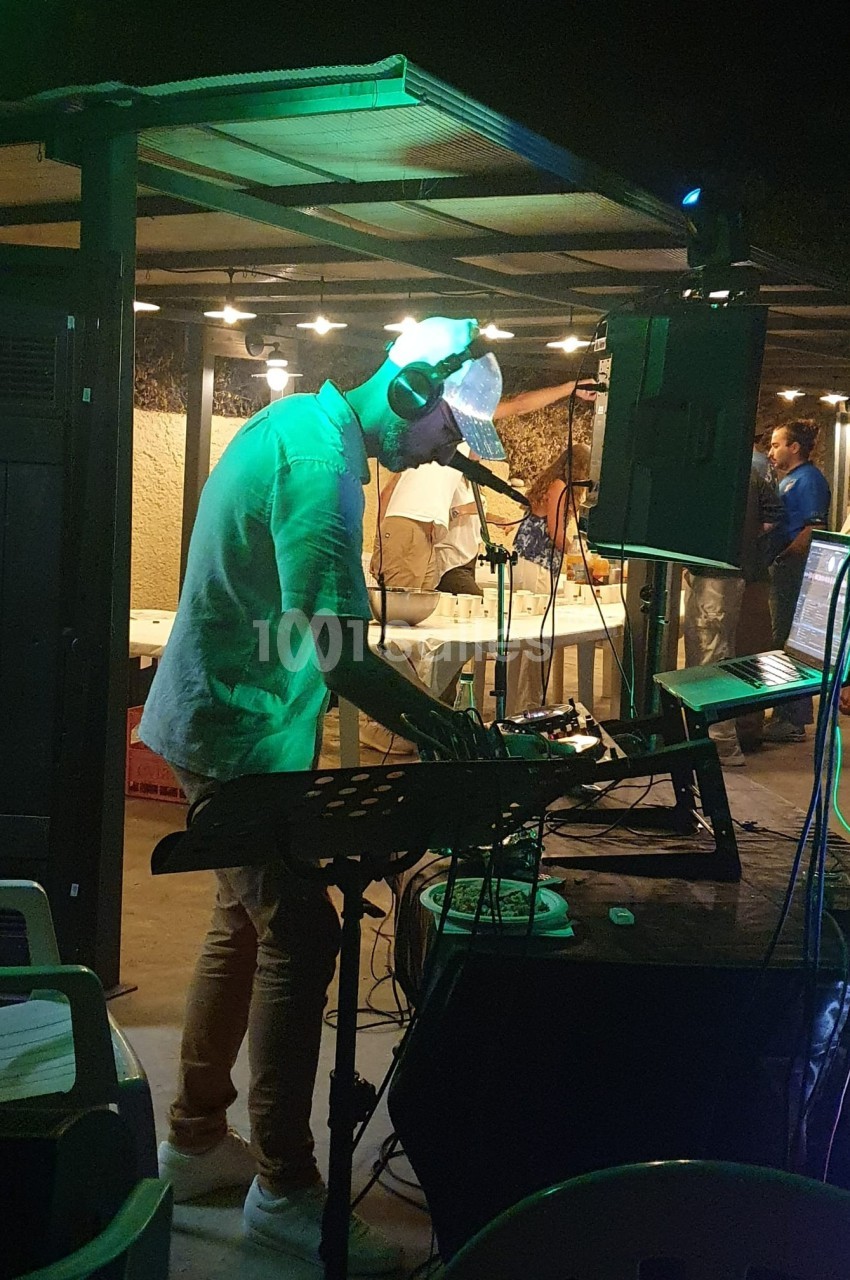 Un DJ mixe de la musique en extérieur, sous un éclairage tamisé, avec des invités en arrière-plan.