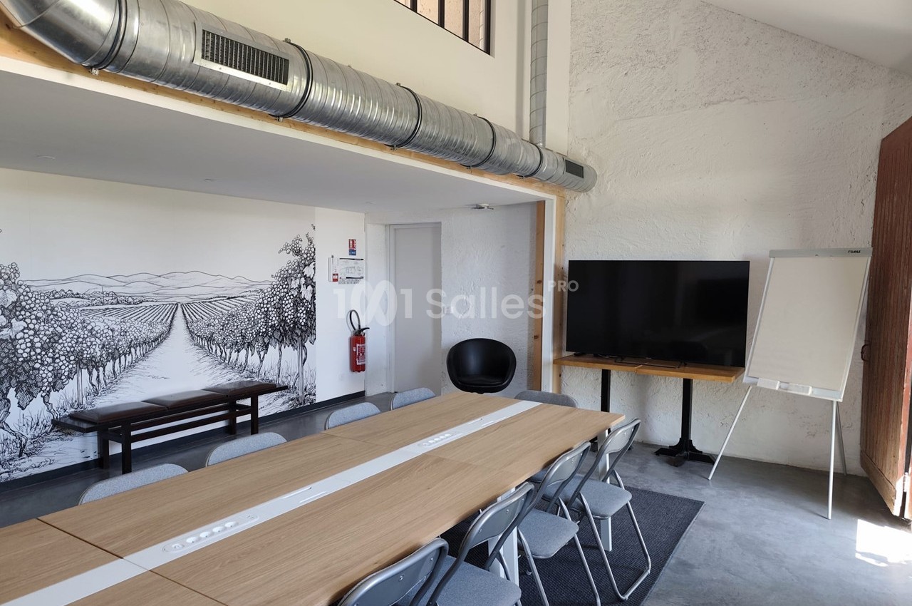 Salle de réunion lumineuse avec table en bois, chaises pliantes, écran, paperboard et fresque murale représentant des vignes.