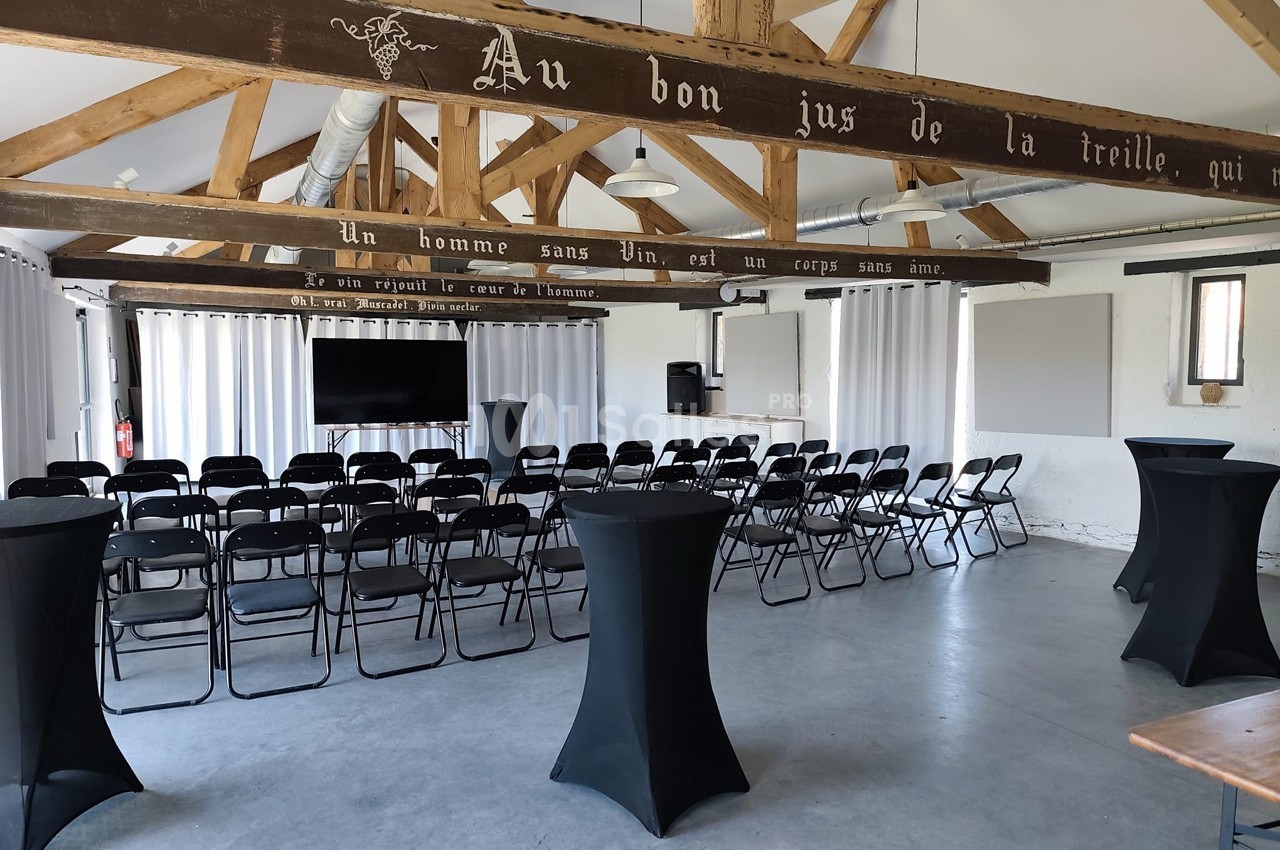 Salle de réunion lumineuse avec chaises alignées, tables hautes noires et poutres apparentes en bois.