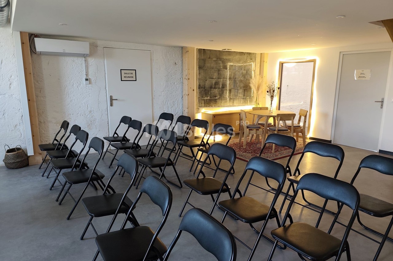 Salle lumineuse avec des chaises pliantes noires disposées en rangées face à une table et un miroir éclairé.