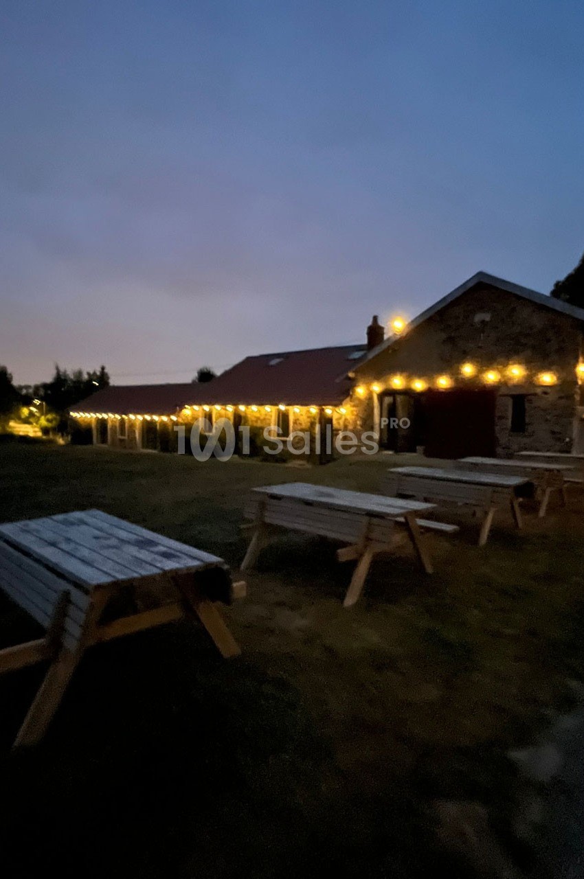 Bâtiment en pierre éclairé par des guirlandes lumineuses au crépuscule, avec des tables en bois sur une pelouse.