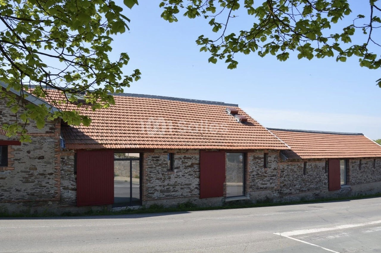 Bâtiment en pierre avec toit en tuiles rouges, grandes portes rouges et fenêtres, bordé par une route et des arbres.