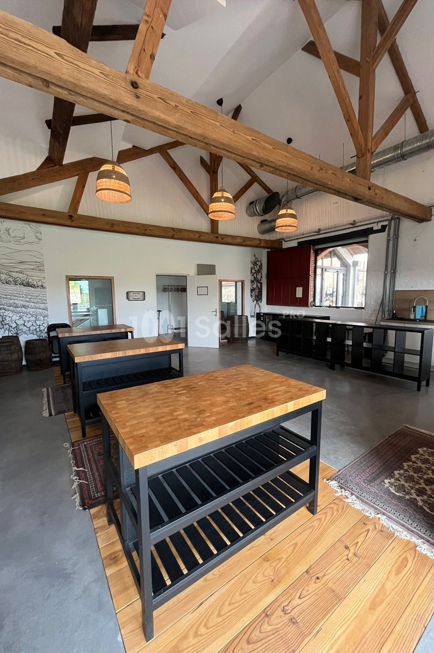 Salle lumineuse avec poutres apparentes, tables en bois et étagères noires, décorée de tapis et éléments rustiques.