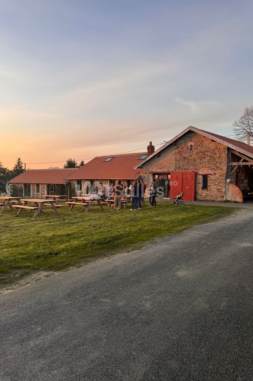 Groupe de personnes discutant devant un bâtiment en pierre avec toit en tuiles, tables de pique-nique et pelouse au coucher…