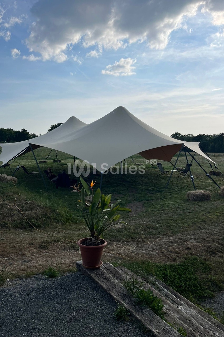 Tente blanche tendue sur une pelouse, entourée de bottes de foin, avec un pot de fleurs au premier plan.