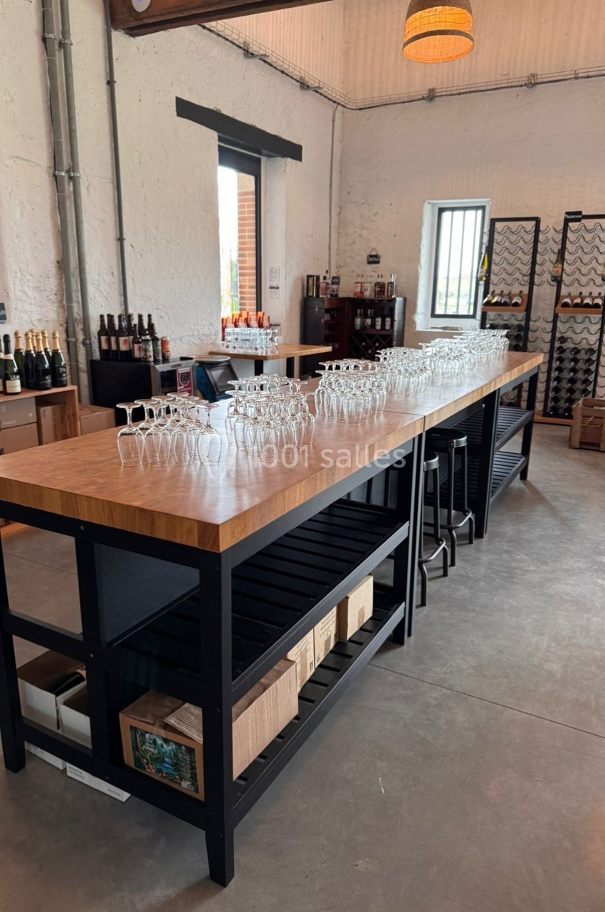 Table en bois avec verres alignés dans une salle lumineuse, entourée de bouteilles et de casiers à vin.