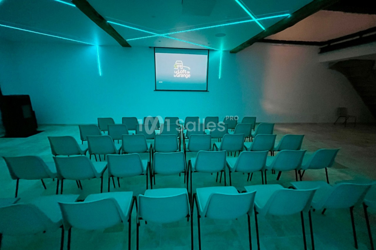 Salle de conférence moderne avec rangées de chaises blanches face à un écran de projection allumé.
