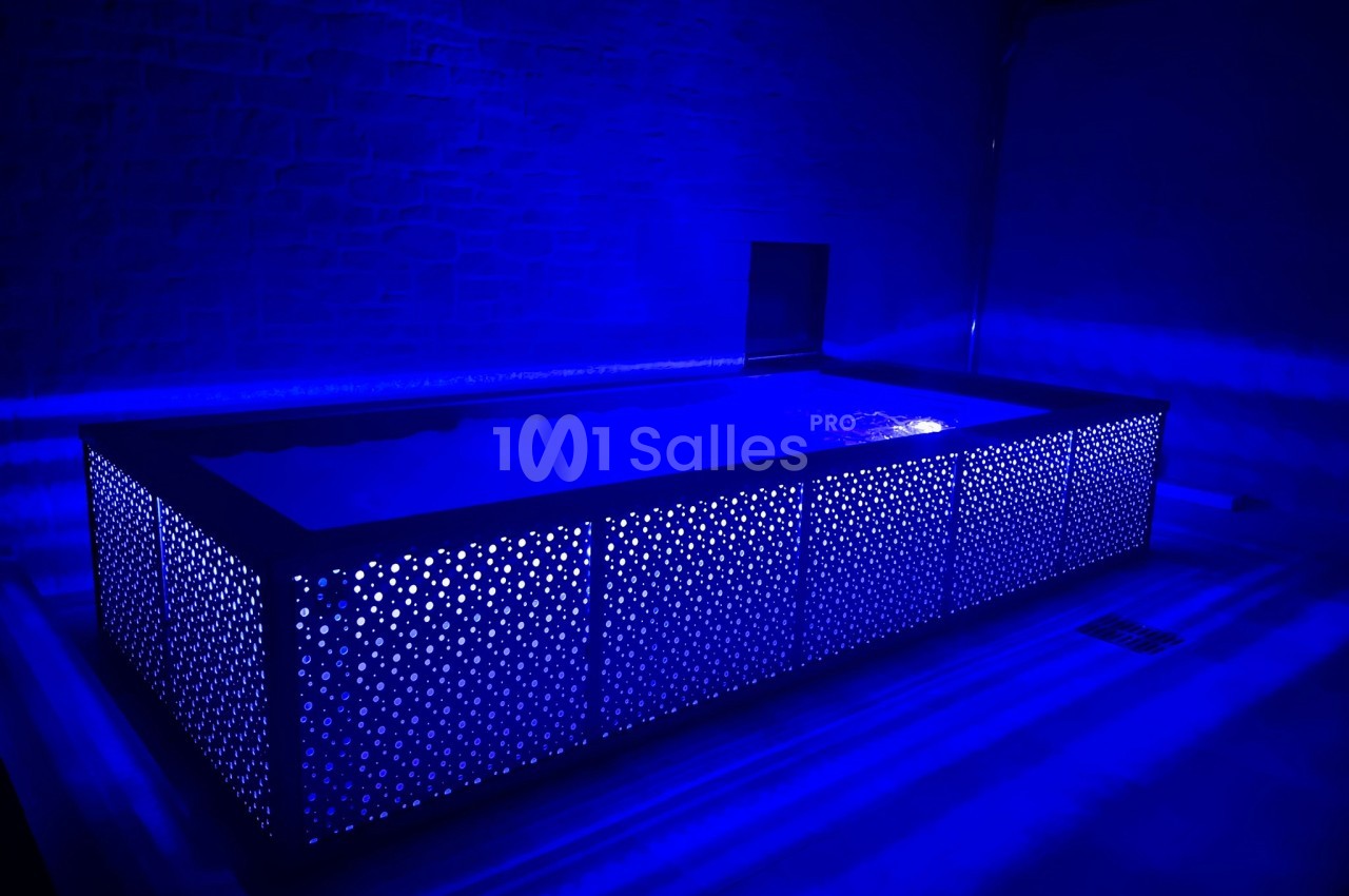 Piscine rectangulaire illuminée par une lumière bleue, entourée d'un panneau perforé dans un espace sombre.